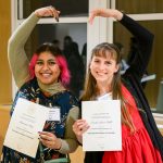Two graduate student women make a heart with their arms while holding their Presidential Fellowship certificates.
