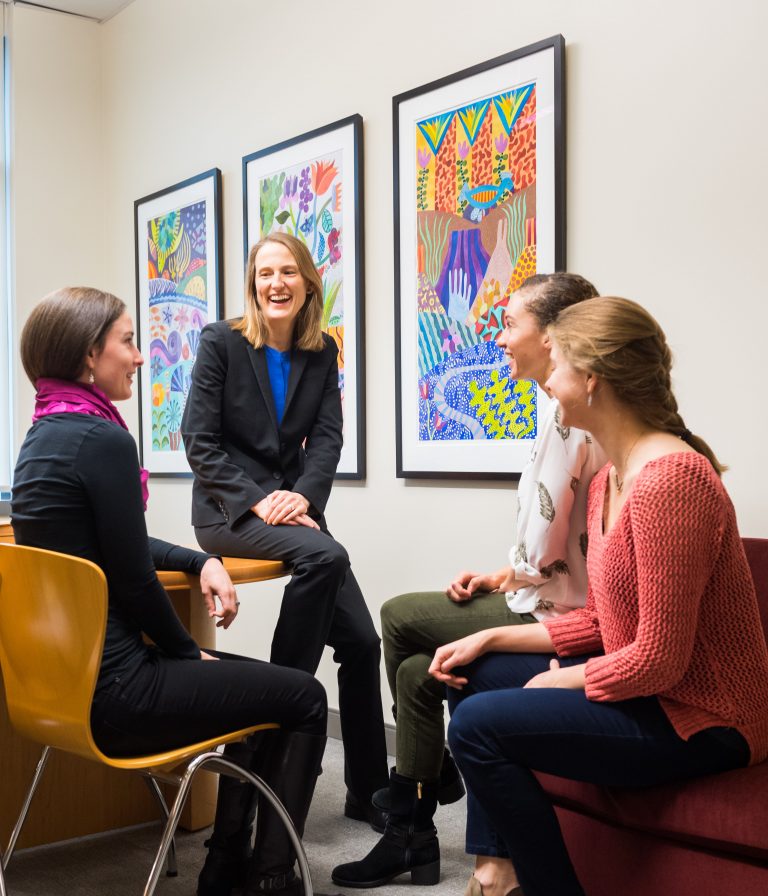 Kate Kellogg laughs with a group of graduate students, sitting in front of colorful paintings.