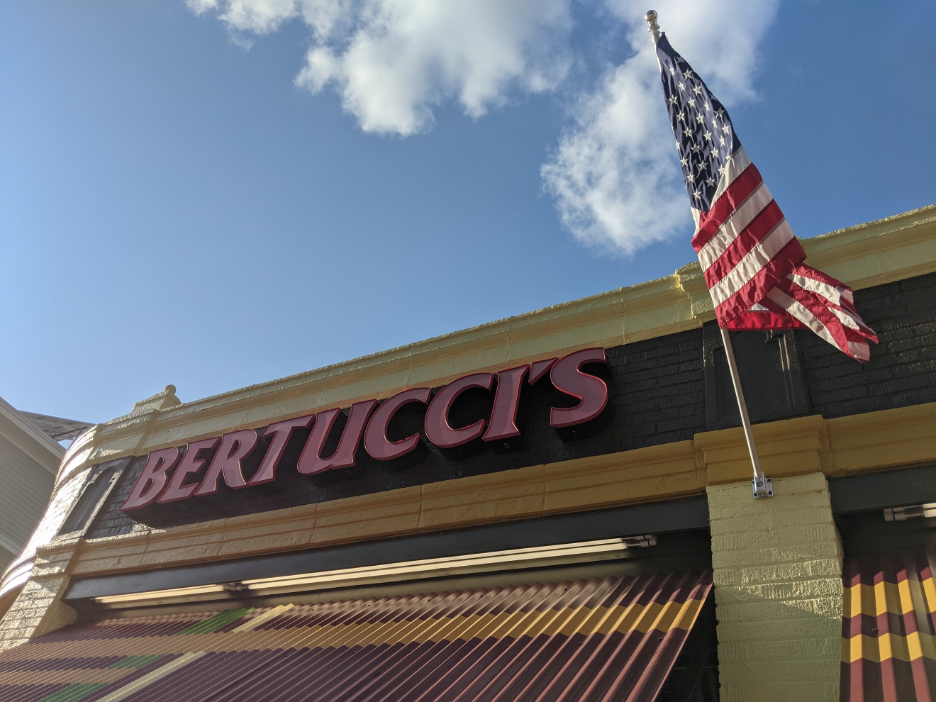 A close-up photo the Bertucci's sign at the top of their building. An American flag is blowing in the wind beside it.