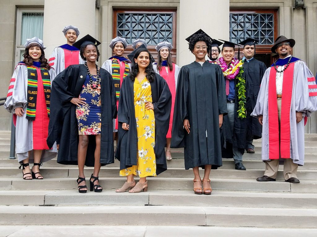 A group of smiling MIT graduate students of color pose together for a photo, wearing graduation gowns and caps.
