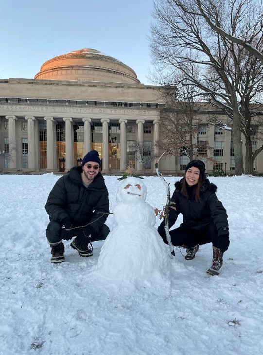 Lizbeth and Lizbeth's partner pose, smiling, with the snowman that they built in front of the MIT Dome. The snowman has leaves for eyes and a hat.