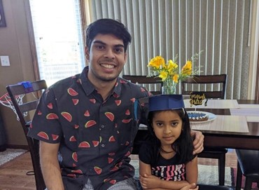 Chinmay sits with his cousin at her graduation party. She is wearing a paper graduation cap and a bouquet of flowers sit on the table behind them.