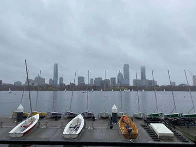 Several sailboats sit on the dock in the foreground of the image; more are seen in the distance on the Charles River. Across the river, clouds obscure the tips of the Boston skyline.