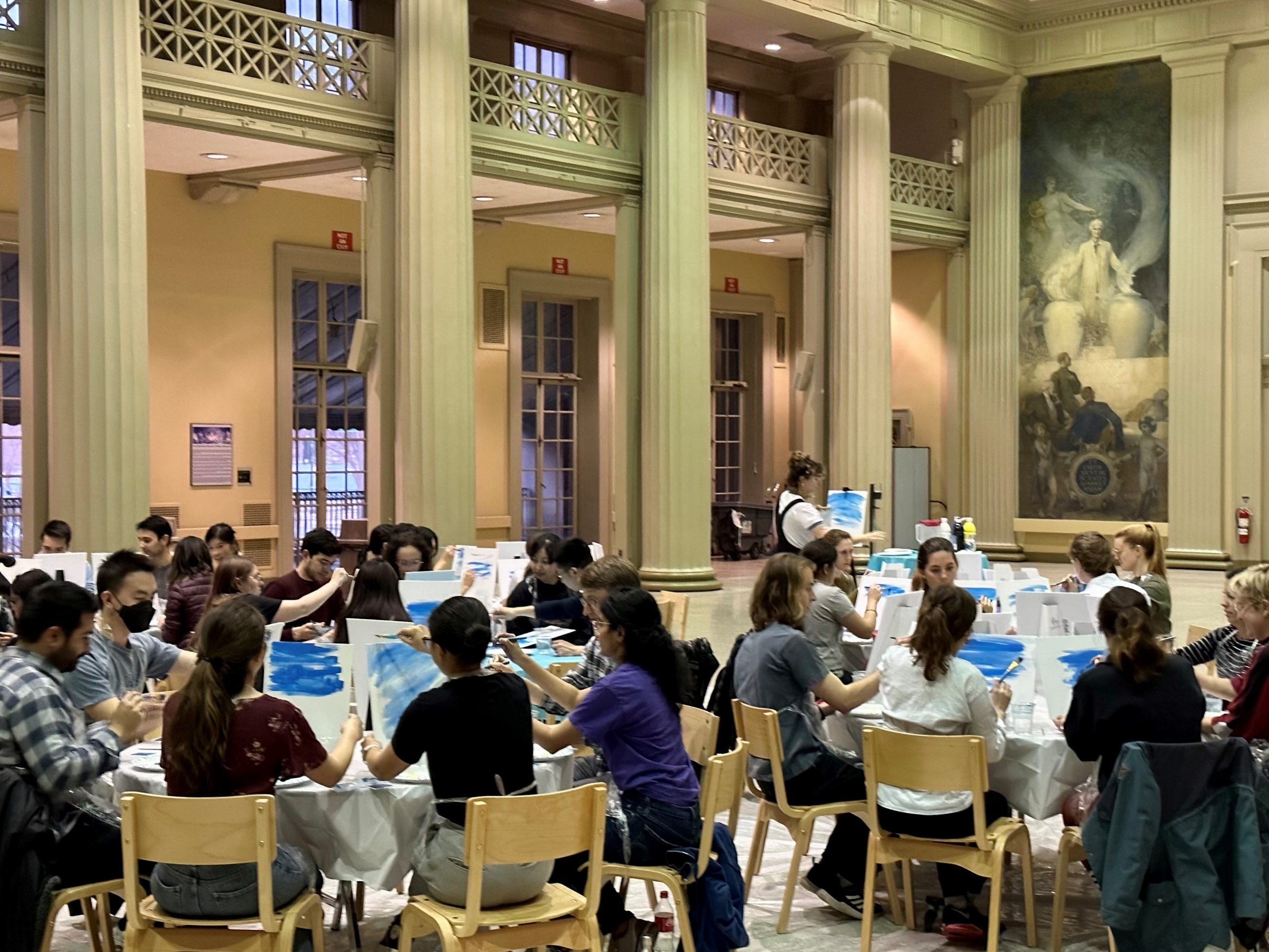 A group of students at round tables paint along with an artist at the front of the room. They're in Morss Hall, a room with beautiful columns and murals on campus.