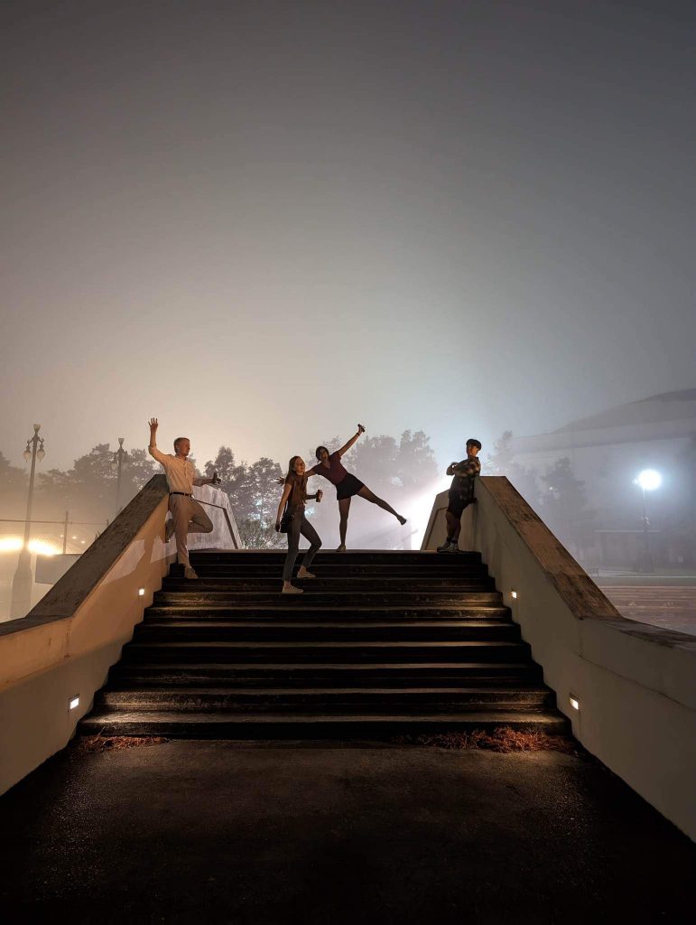 On a foggy night, a group of people strike big poses at the top of an outside stairway.