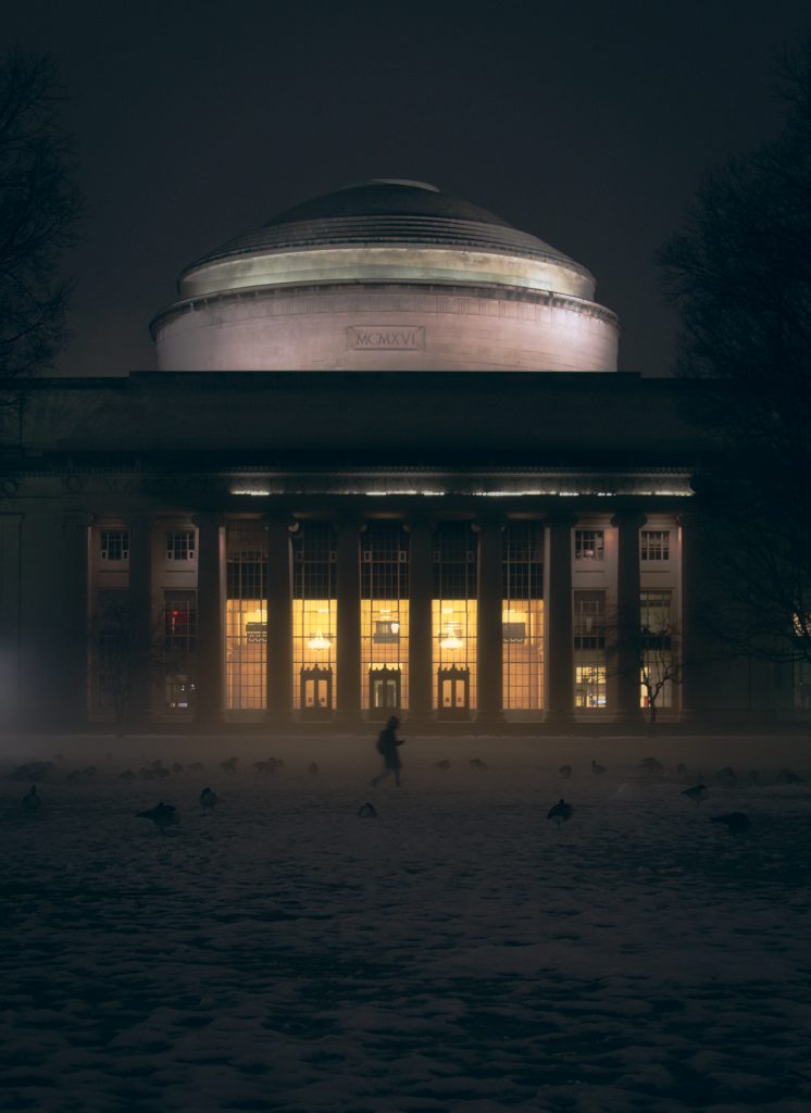 The MIT dome and Lobby 10 take up most of the photo, seen from Killian Court at night; the warm lights glow through the lobby windows as a lone figure walks across the court in the misty darkness. Geese are scattered throughout the lawn.