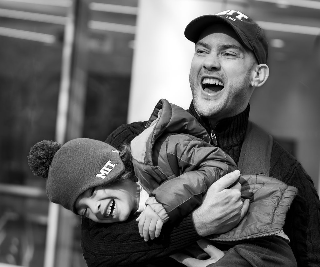 A male graduate student holds his young child in a playful pose. Both are laughing and wearing MIT hats.