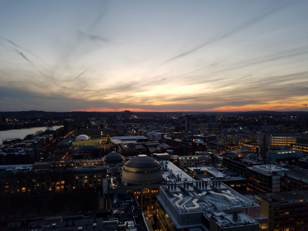 Aerial view of MIT's campus, with the dome at center. The sun has already set, and there is a warm orange glow and scattered clouds on the horizon. Many windows are lit. 