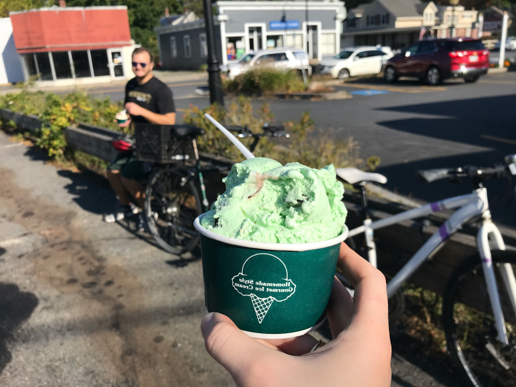 Miriam holds up a cup of green ice cream in front of a sunny, green-lined parking lot.