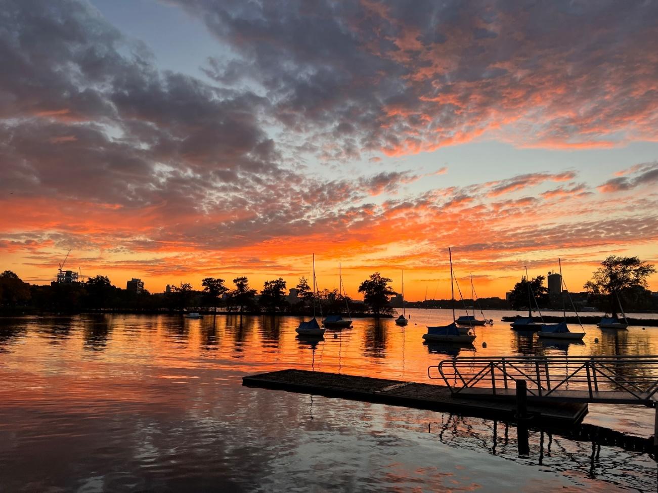 A glorious, vibrant sunset reflected on the Charles River. Silhouettes of docked boats and leafy trees rim the horizon.