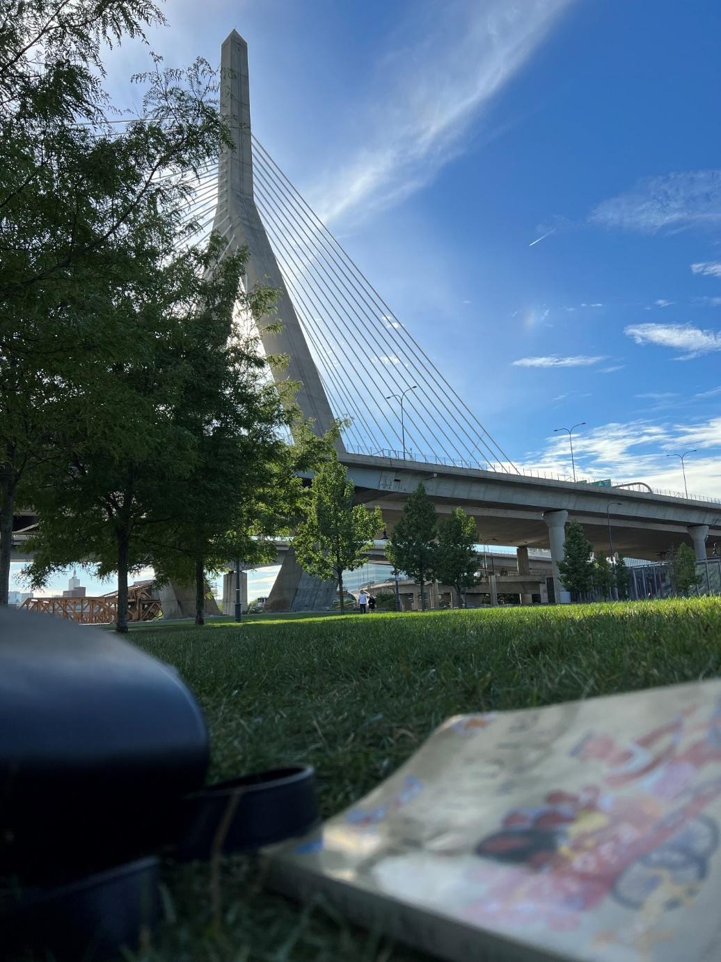 Shot from low to the ground, a blurred book sits in the foreground while the camera focuses on Boston's iconic Zakim Bridge on a sunny day.