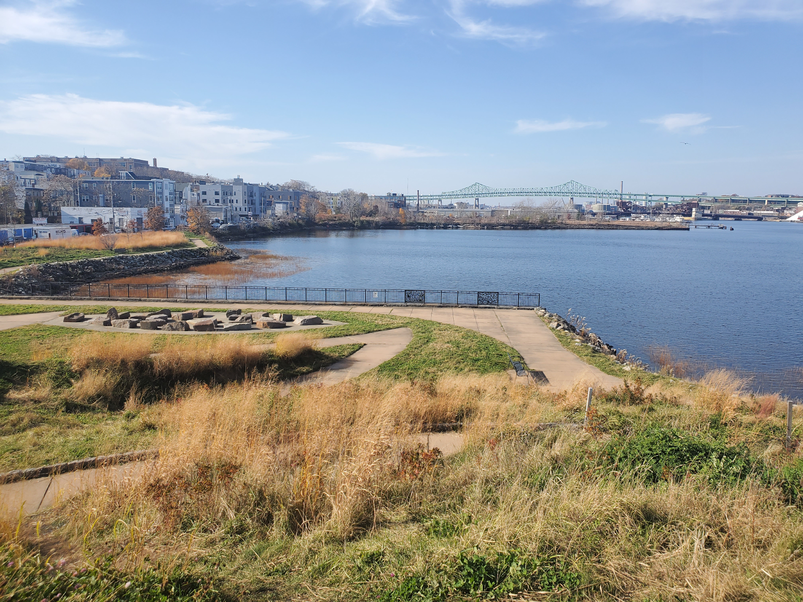 Sunshine lights a waterfront view with apartment buildings and a bridge in the background and a charming grassy park in the foreground.