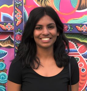 A smiling dark haired graduate student stands against a background of vibrantly colored artwork