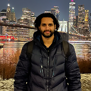 A smiling dark haired man stands in front of a city skyline