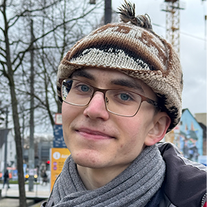 A smiling graduate student in glasses and a hat stands outdoors
