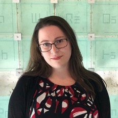 A dark haired woman with glasses stands in front of a wall with artful glass squares representing each periodic element