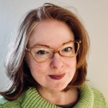 A red haired, bespectacled woman smiles at the camera