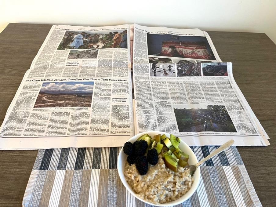 An open newspaper on a table, with a bowl of food sitting in front of it on a blue and brown placemat