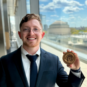 A graduate student stands by a window and smiles, wearing glasses and a navy suit and tie and holding his Goodwin Medal