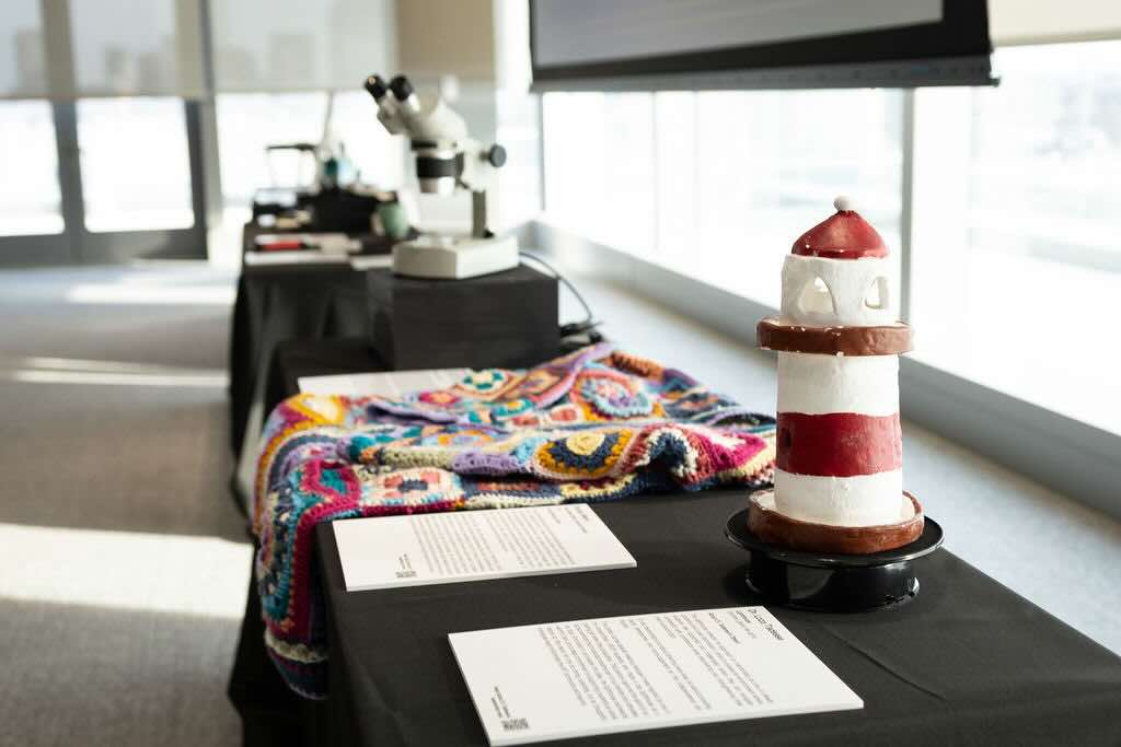 A handmade lighthouse sits atop a black table, with a multicolored crocheted shawl in the background. A microscope sits on another table further in the background.