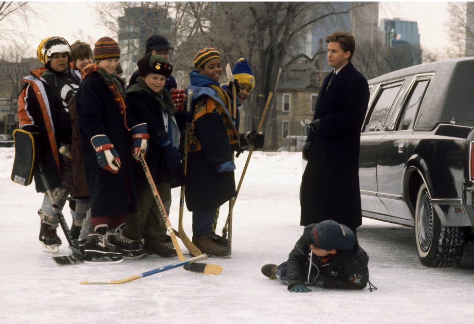 This scene from the Mighty Ducks shows a huddle of young boys with hockey sticks, wearing hats and winter coats, facing a well dressed man in a long coat.