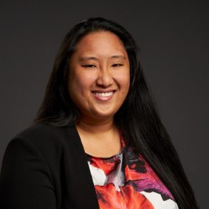 A brown haired woman smiles at the camera, wearing a floral blouse and black blazer.