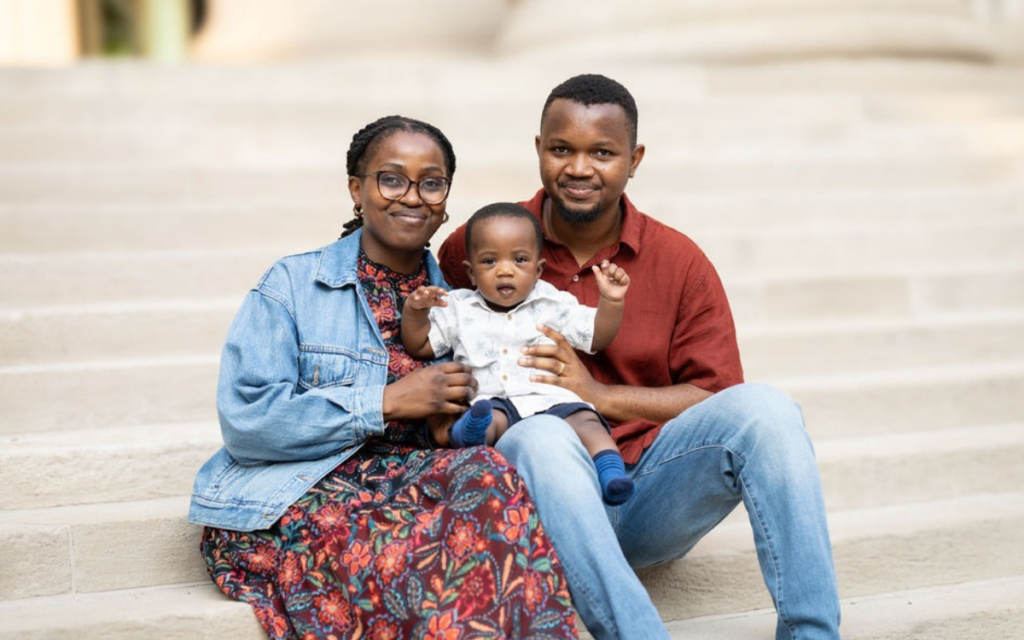A female graduate student poses on the steps of 77 Mass Ave., holding her baby alongside her husband